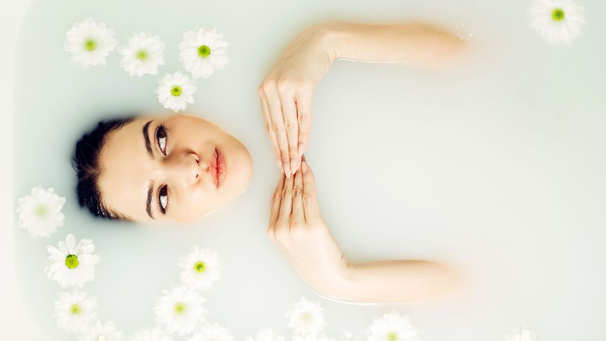 Happy young woman is lying in a milk bath with flowers, top view