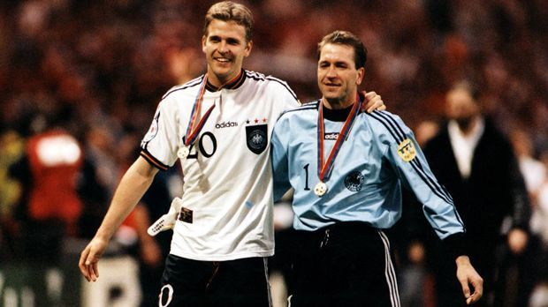 
                <strong>EM 1996 (Oliver Bierhoff und Thomas Köpke)</strong><br>
                EM 1996: In England holen Oliver Bierhoff (li.), Thomas Köpke und Co. den zweiten EM-Titel für Deutschland. Im Finale schlägt die Mannschaft in den schlichten weißen Trikots das tschechische Nationalteam. Neu an den Leibchen ist das schwarz umrandete Wappen auf der Brust und die drei Sterne, für die drei gewonnenen WM-Titel.
              