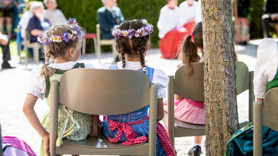 Drei Mädchen in Tracht sehen sich einen Festgottesdienst zu Fronleichnam unter freiem Himmel am bayerischen Staffelsee an.

