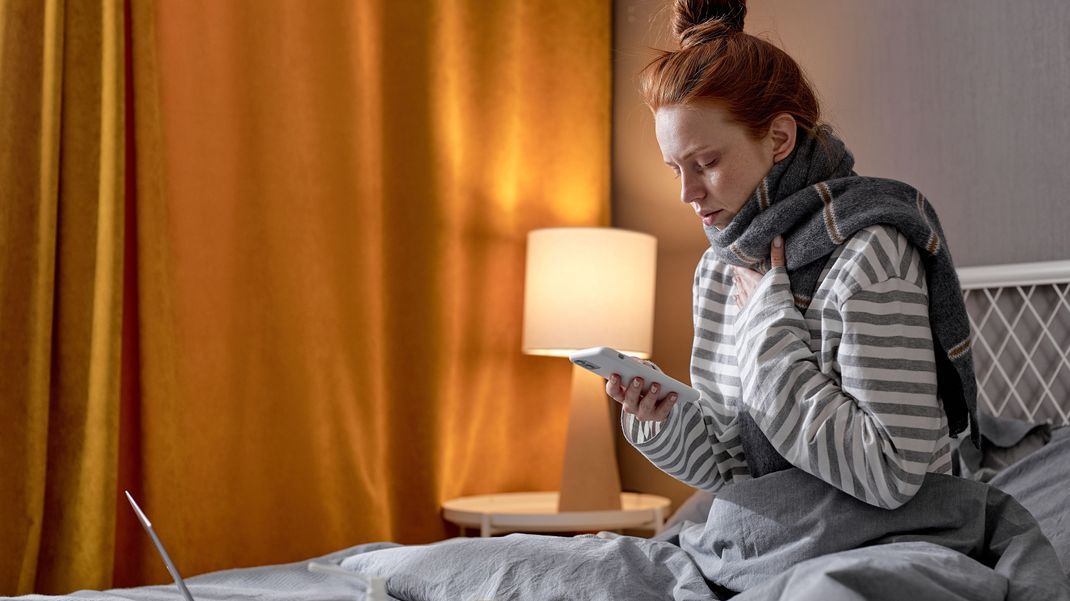 Eine Frau sitzt krank im Bett und - nutzt die Option der telefonischen Krankmeldung.