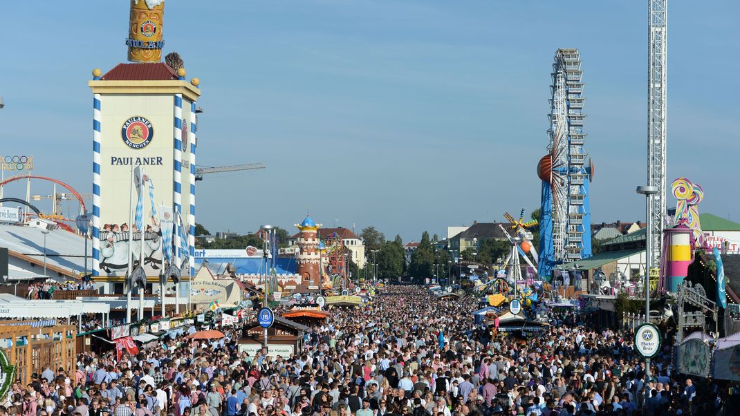 Zahlreiche Menschen gehen bei Sonnenschein über das Oktoberfest-Gelände in München.