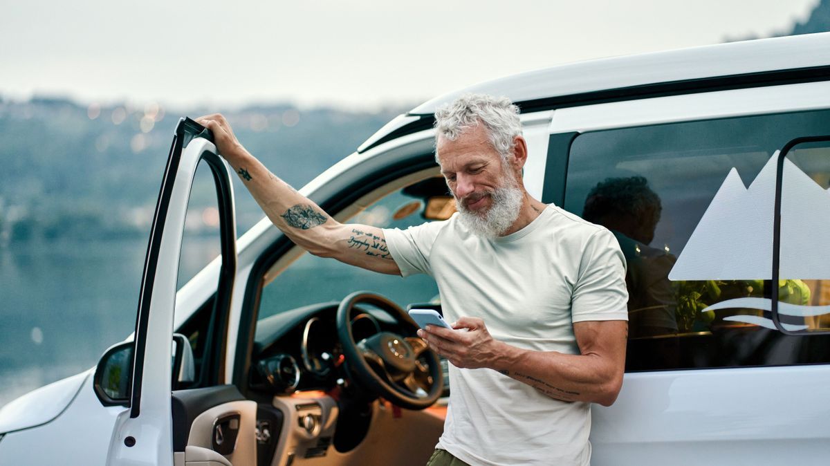 Mature man traveler standing near camper van using mobile in camping.
