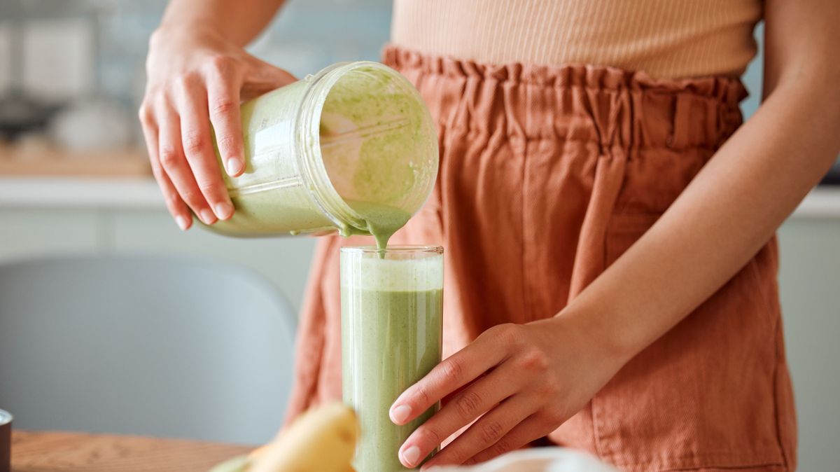 Woman pouring healthy smoothie in a glass from a blender jar on a counter for detox. Female making fresh green fruit juice in her kitchen with vegetables and consumables for a fit lifestyle.