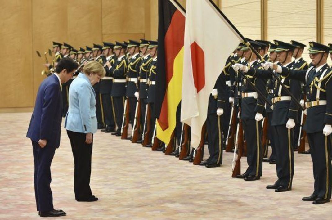 Die ehemalige deutsche Bundeskanzlerin und der japanische Premierminister verbeugen sich vor einer Ehrengarde
