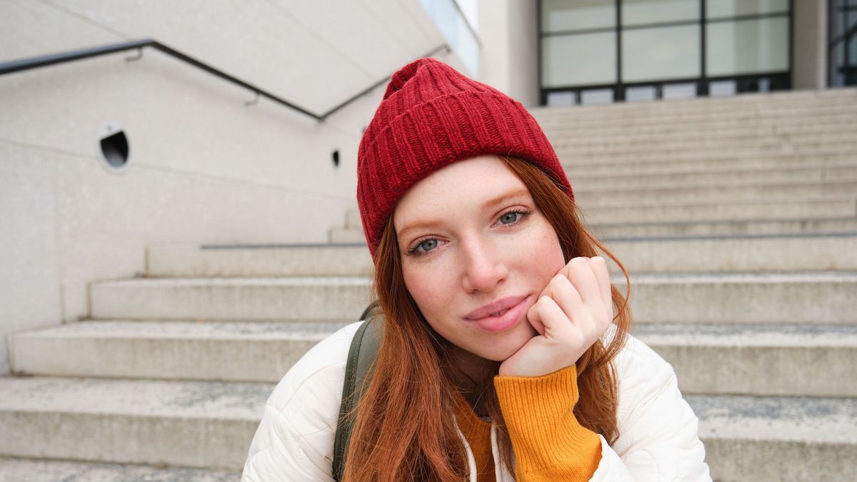 Close up portrait of beautiful redhead girl in red hat, urban woman with freckles and ginger hair, sits on stairs on street, smiles and looks gorgeous