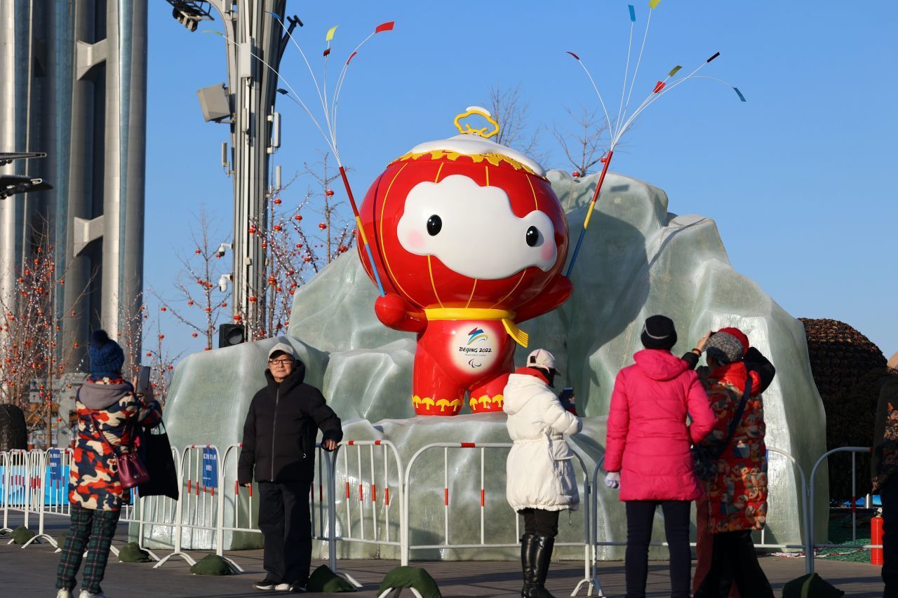 Das Maskottchen: Shuey Rhon Rhon ist ein Laternenkind. Traditioneller chinesischer Papierschnitt inspirierte das Design. Sein Name steht für die Inklusion von Menschen mit Handicap in der Bevölkerung. „Shuey“ bedeutet in diesem Zusammenhang Schnee und das erste „Rhon“ so viel wie einbeziehen und tolerieren. Das zweite „Rhon“ bedeutet warm und verschmelzen. Sein leuchtendes Herz zeigt das Logo der Paralympischen Winterspiele. 