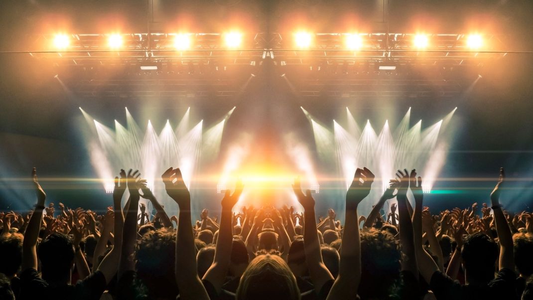 Photo of a concert hall with people silhouettes clapping in front of a big stage lit by spotlights. Shot is taken from concert crowd point of view, lens flare is visible.