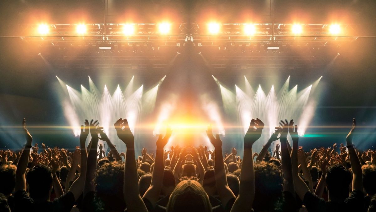 Photo of a concert hall with people silhouettes clapping in front of a big stage lit by spotlights. Shot is taken from concert crowd point of view, lens flare is visible.