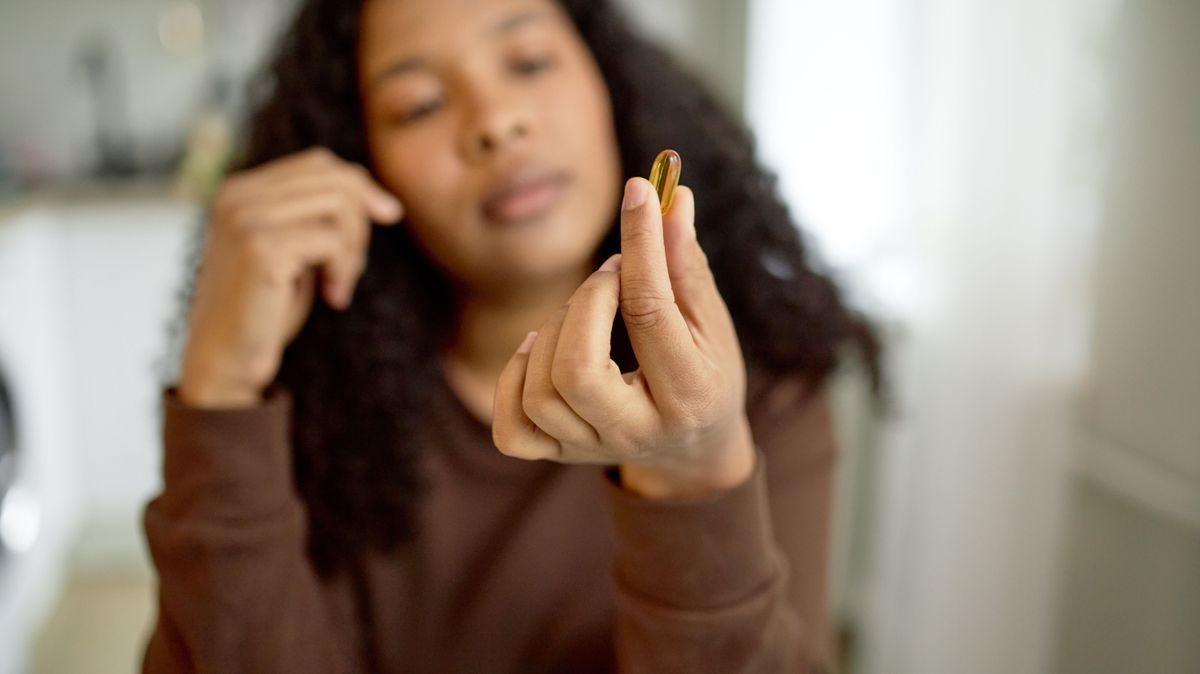 Selective focus on hand with transparent yellow vitamin pill of polyunsaturated fat acids or omega-3 fish oil, blurred puzzled and thoughtful face expression of African American female on background