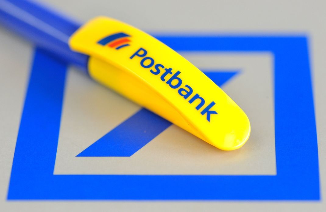 Auf dem Logo der Deutschen Bank liegt ein Kugelschreiber mit der Aufschrift "Postbank".