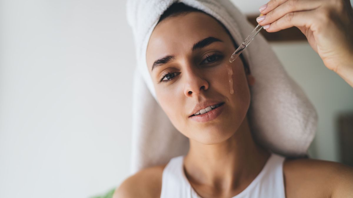 Young female with towel on head puts moisturizing serum on face while standing at bathroom. 30 years old woman doing daily morning rituals. Enjoying healthy skin with collagen care`, skincare product