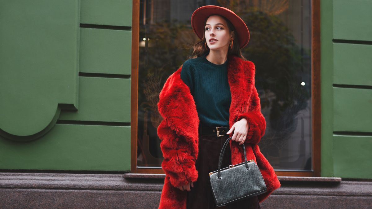  Outdoor fashion portrait of young beautiful confident lady wearing trendy orange faux fur coat, hat, green sweater, holding stylish snakeskin textured handbag, posing in street of city. Copy space