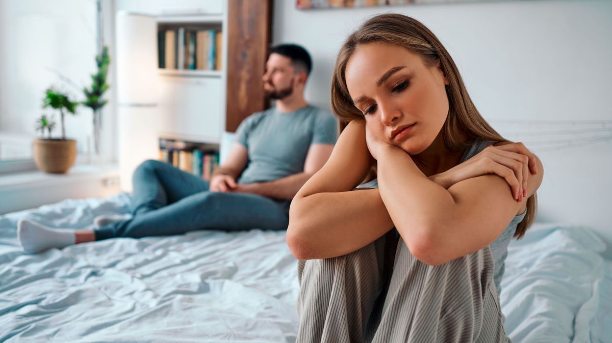 Stashing-Dating Trend-Upset young woman in home clothes sitting on the bed, in the background sitting man. Family troubles.