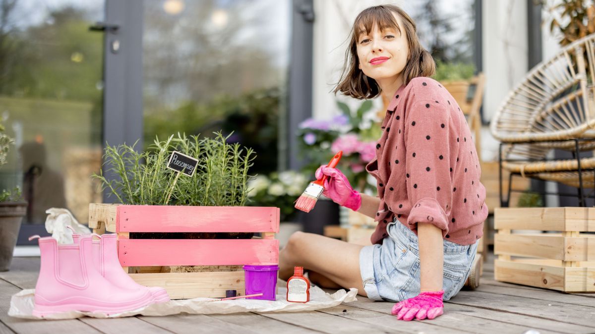 Young woman painting wooden box in pink color, doing some renovating housework on the terrace outdoors. DIY concept