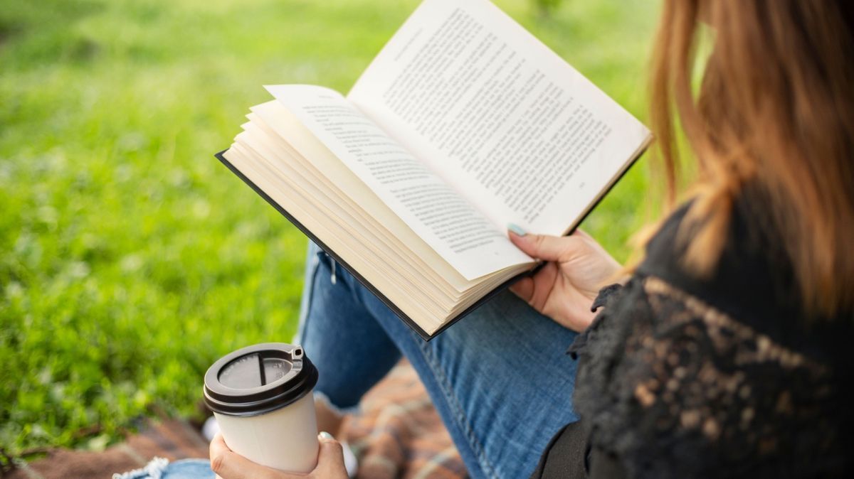A woman sits near a tree in the park and holds a book and a cup with a hot drink in her hands. A woman in jeans and a t-shirt reading a book outdoor