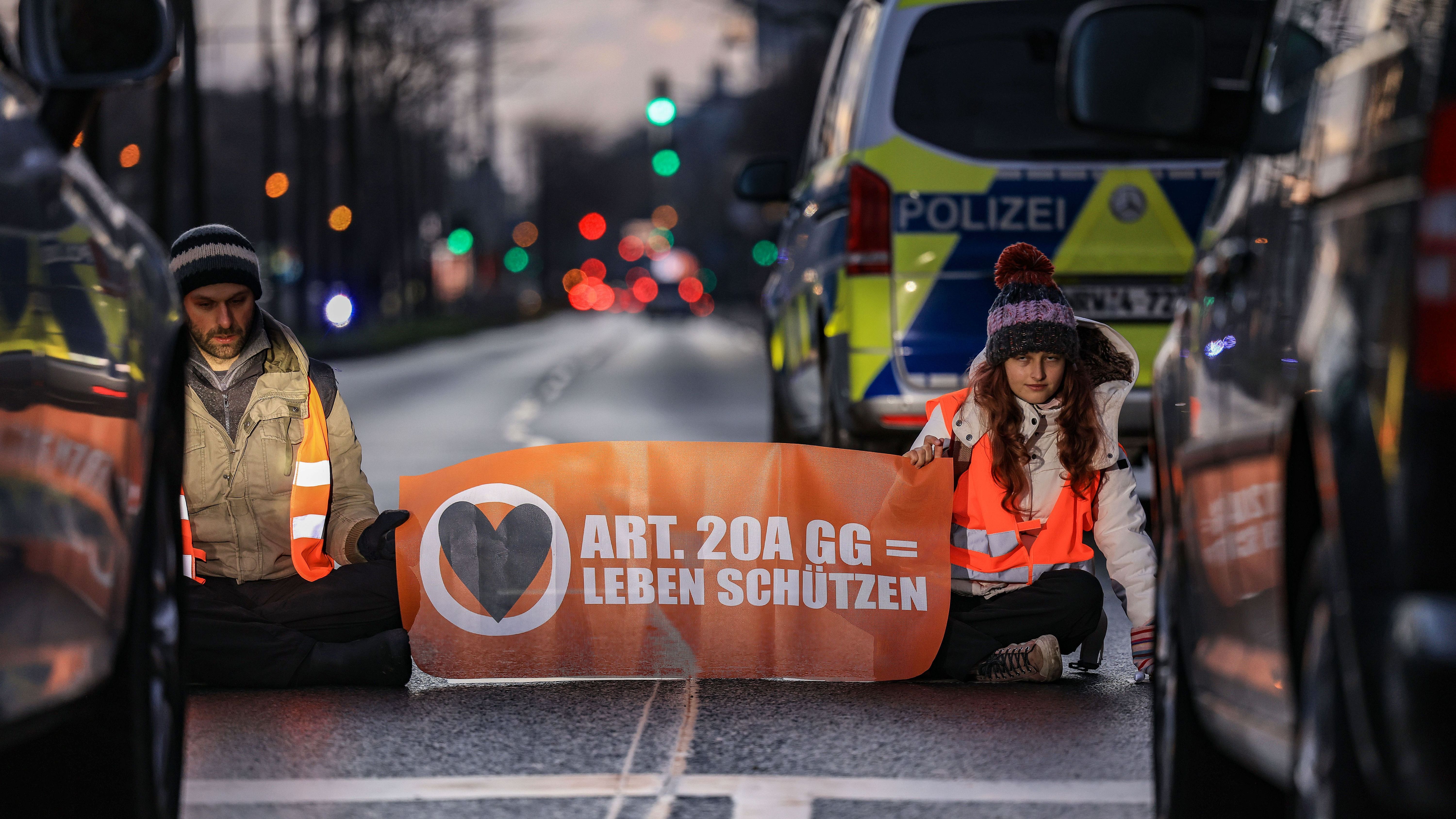 Aktivist:innen der Umweltschutzorganisation "Letzte Generation" sitzen vor Fahrzeugen auf einer Straße.