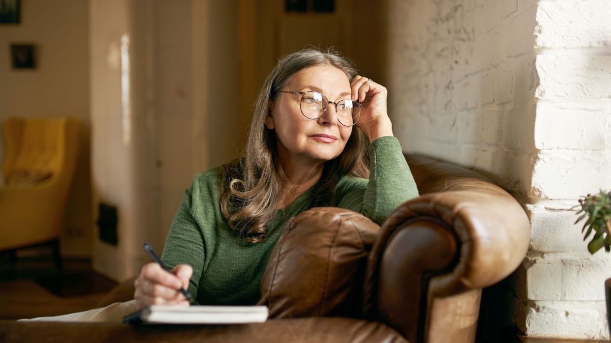 People, age, retirement and occupation concept. Attractive thoughtful retired female in stylish round glasses working from home sitting on sofa looking through window, holding pen, making notes