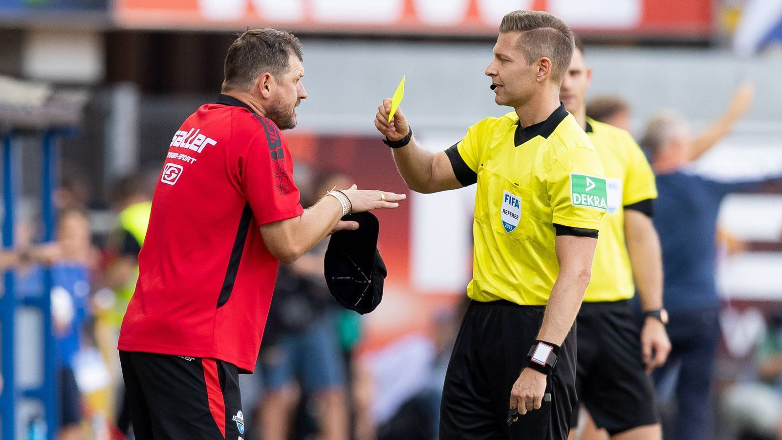 
                <strong>Steffen Baumgart sieht erste Gelbe Karte </strong><br>
                Eine neue Regelung sorgte am 2. Spieltag der Bundesliga-Saison 2019/20 für ein Novum! Paderborns Trainer Steffen Baumgart (li.) sah bei der 1:3-Niederlage des Aufsteigers gegen den SC Freiburg von Schiedsrichter Tobias Welz (re.) als erster Bundesliga-Coach der Geschichte die Gelbe Karte. Auch am 2. Spieltag, aber zeitlich etwas später als Baumgart, sah noch Fortuna Düsseldorfs Coach Friedhelm Funkel ebenfalls Gelb an der Seitenlinie. Sollte ein Trainer innerhalb einer Spielzeit vier Gelbe Karten kassieren, wird er - genau wie bei den Spielern - für eine Bundesliga-Begegnung gesperrt. 
              