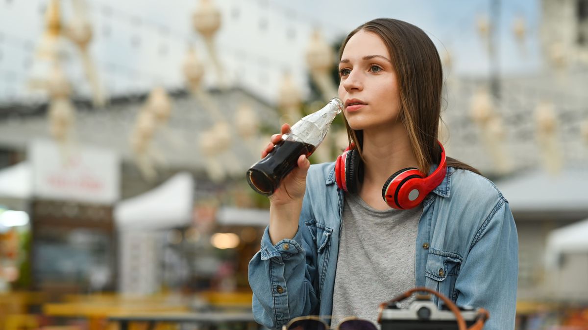Attractive woman with wireless headphone drinking cola from glass bottle while sitting at outdoor street food restaurant