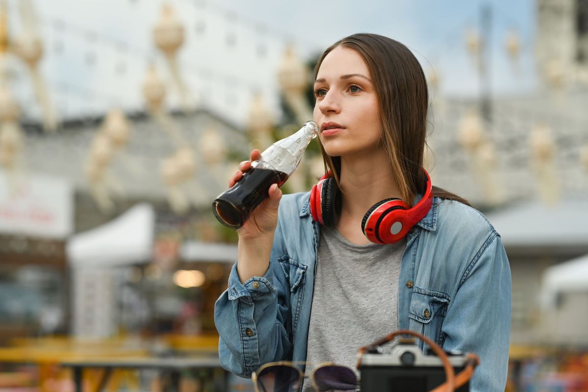 Attractive woman with wireless headphone drinking cola from glass bottle while sitting at outdoor street food restaurant