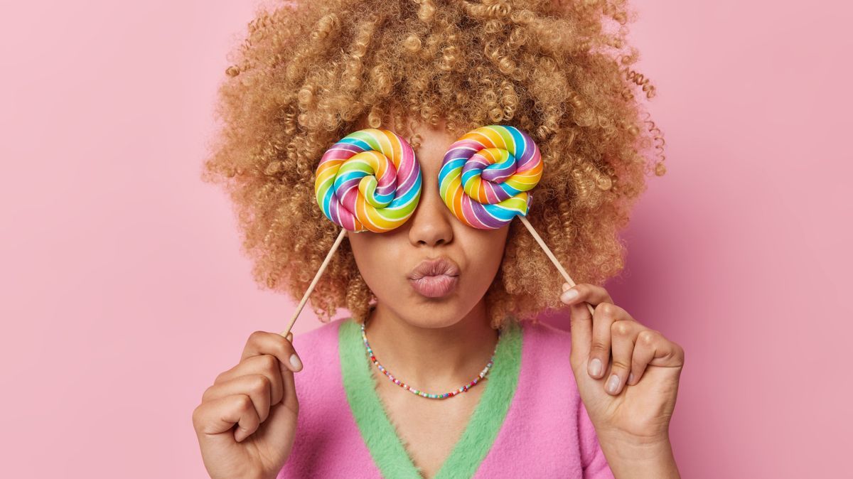 Photo of curly haired young woman covers eyes with rainbow caramel candies keeps lips folded wears casual clothes poses against pink studio background. Sweet tooth and unhealthy nutrition concept