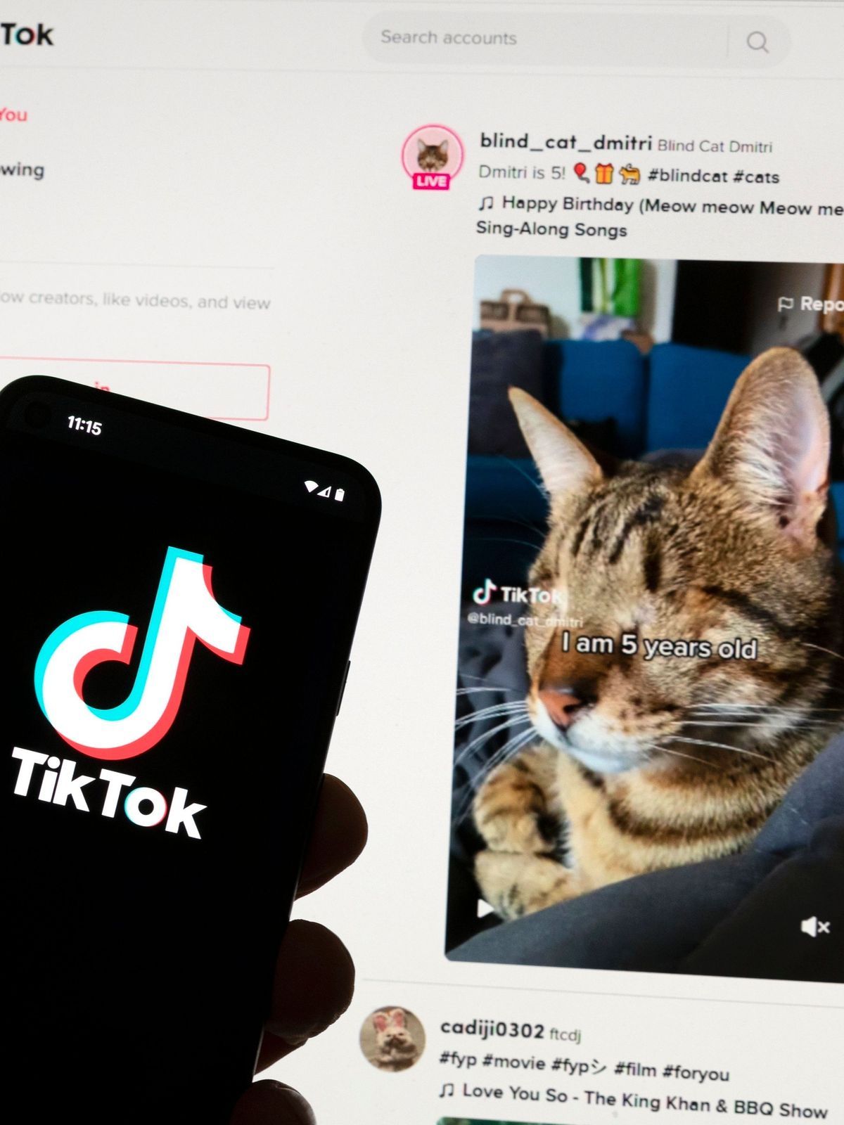 Das TikTok-Logo ist auf einem Mobiltelefon vor einem Computerbildschirm zu sehen, der den TikTok-Startbildschirm anzeigt.