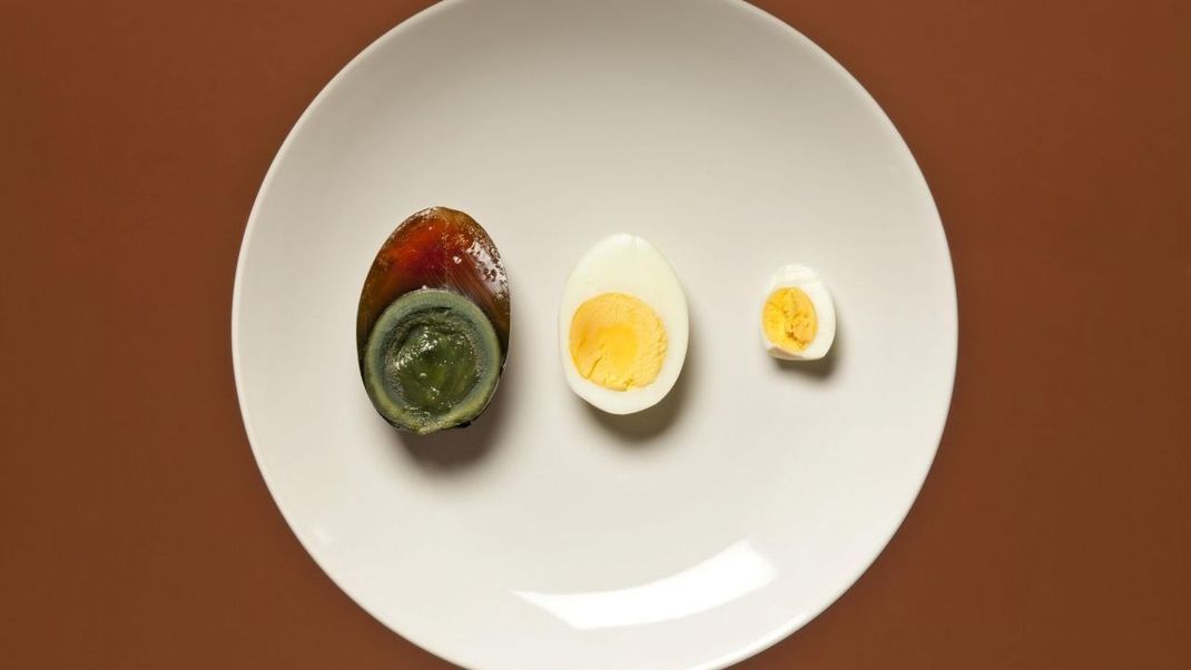 Das Bild zeigt von links nach rechts halbierte Hundertjährige- oder Tausendjährige fermentierte- oder chinesische Eier sowie ein Hühnerei und ein Wachtelei.