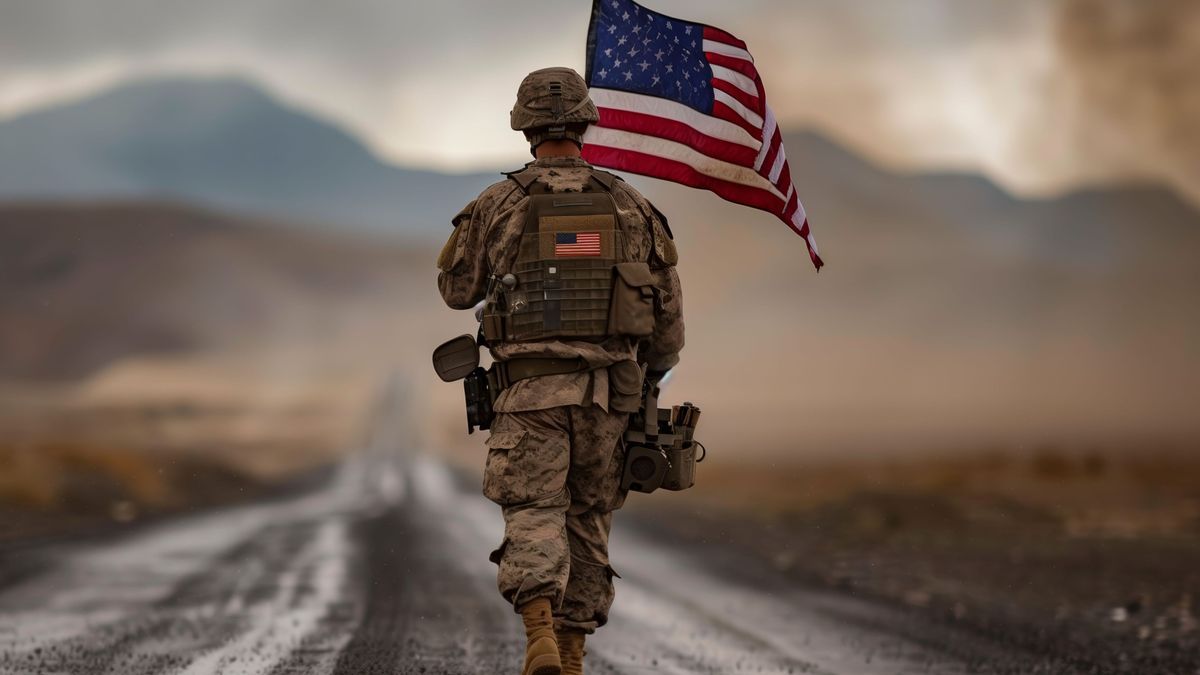 Soldier walks with US flag on road, under sky, in landscape