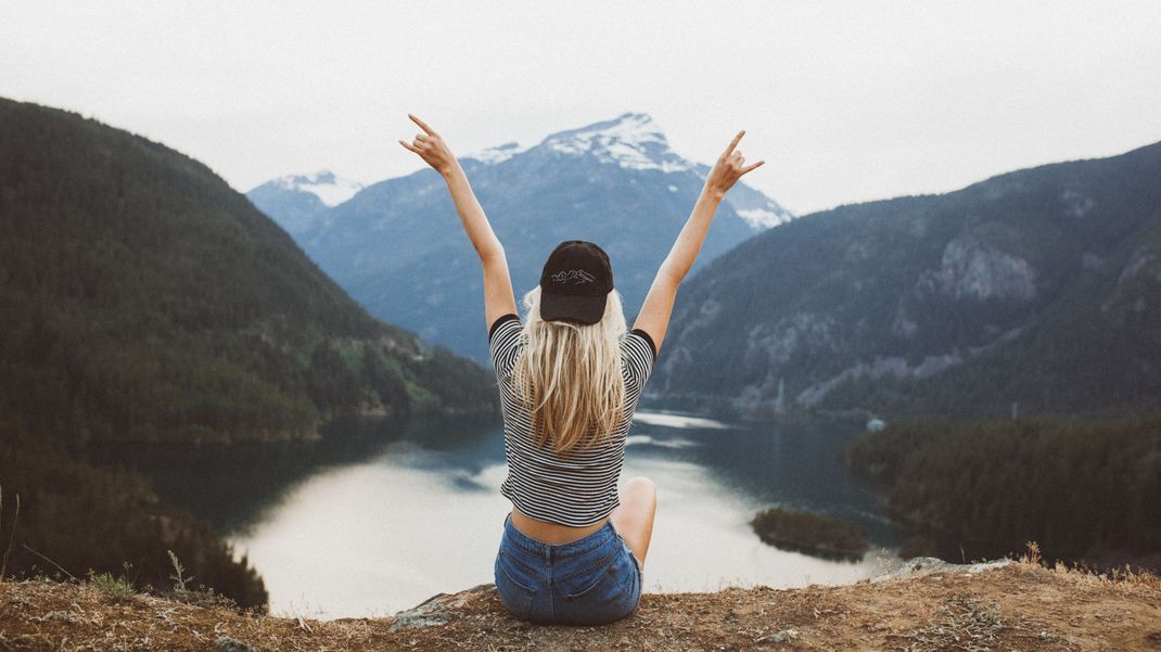 A behind shot of a young blond girl sitting on the cliff enjoying the view of the mountains and lake