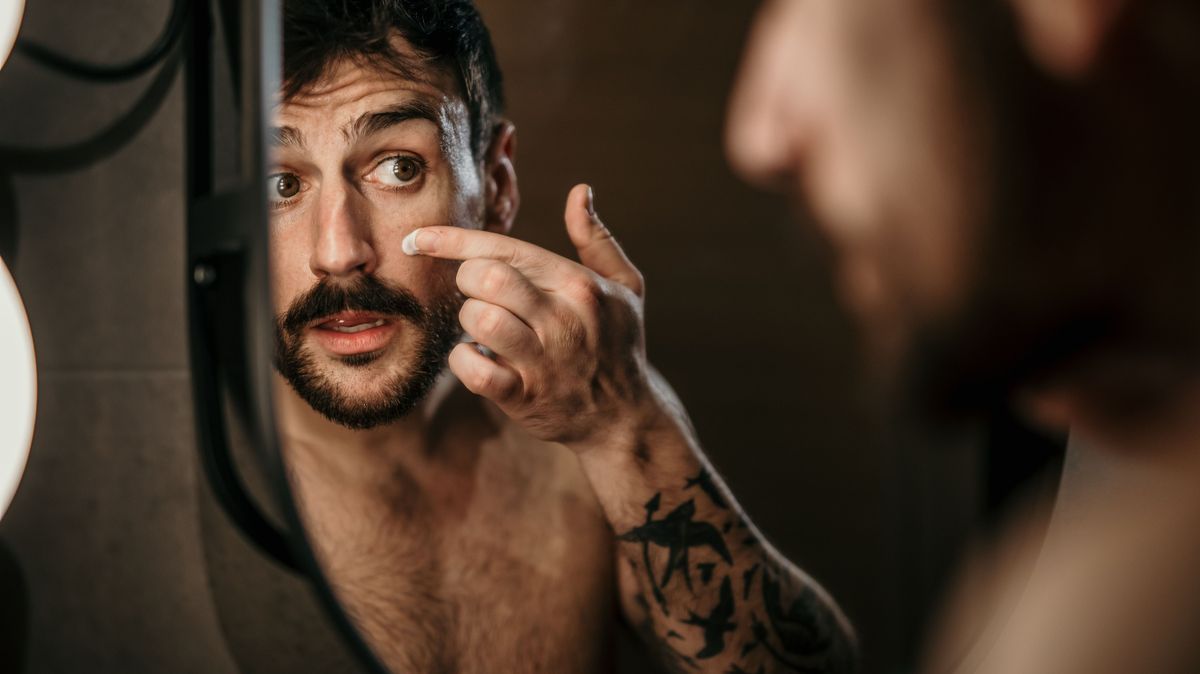 Handsome man looking at himself in the mirror in the bathroom and applying face cream.