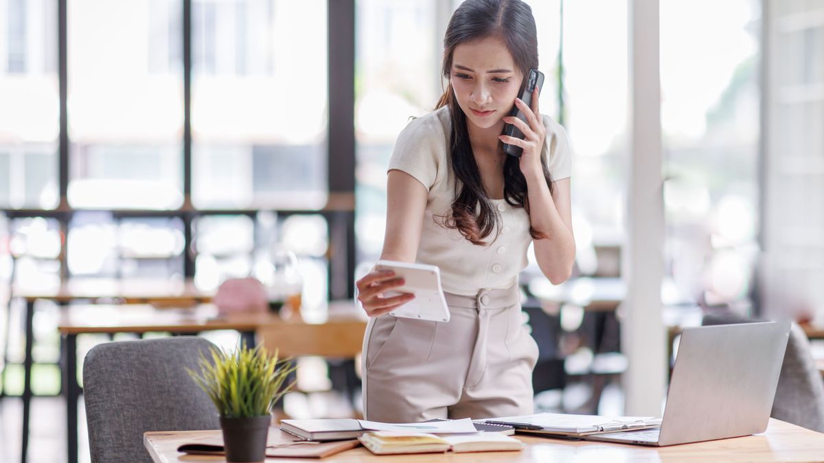 Smiling asian female hold cell at ear talk look at laptop screen. Busy young asian lady advisor lawyer give phone consultation from workplace discuss electronic document details