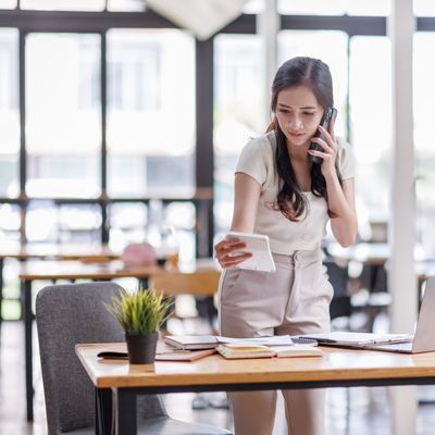 Smiling asian female hold cell at ear talk look at laptop screen. Busy young asian lady advisor lawyer give phone consultation from workplace discuss electronic document details