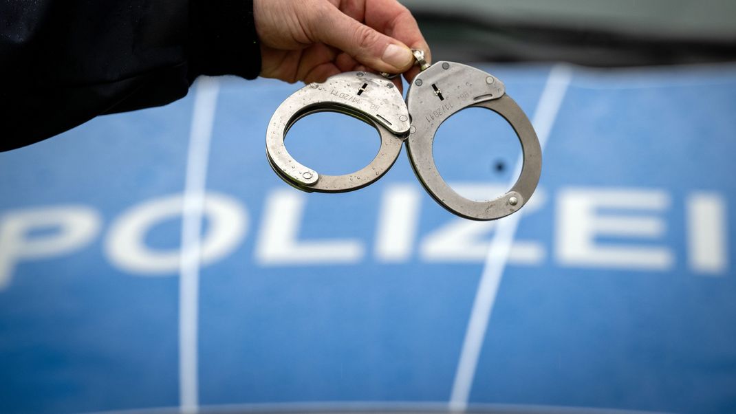 Ein Polizist hält vor einem Streifenwagen Handschellen in der Hand.