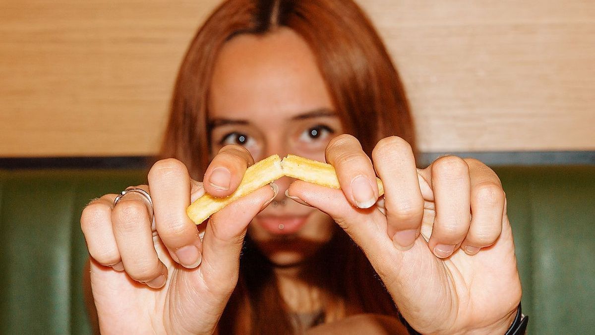 Young woman with red hair and piercing sitting at table and looking at camera while showing french fries in restaurant