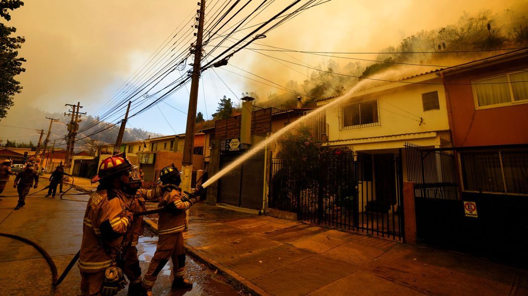 Feuerwehrleute spritzen Wasser auf Häuser, während in der Nähe Waldbrände brennen. 