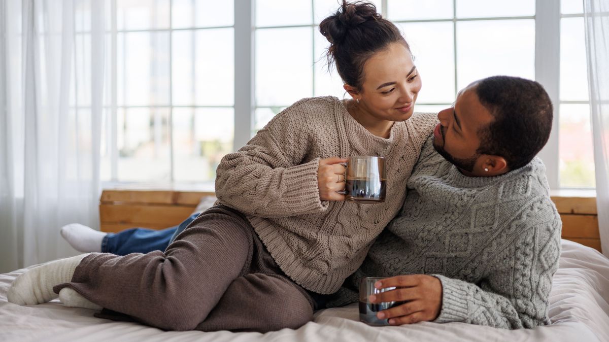 Smiling young asian woman in sweater holding coffee and taking to african american boyfriend on bed at home