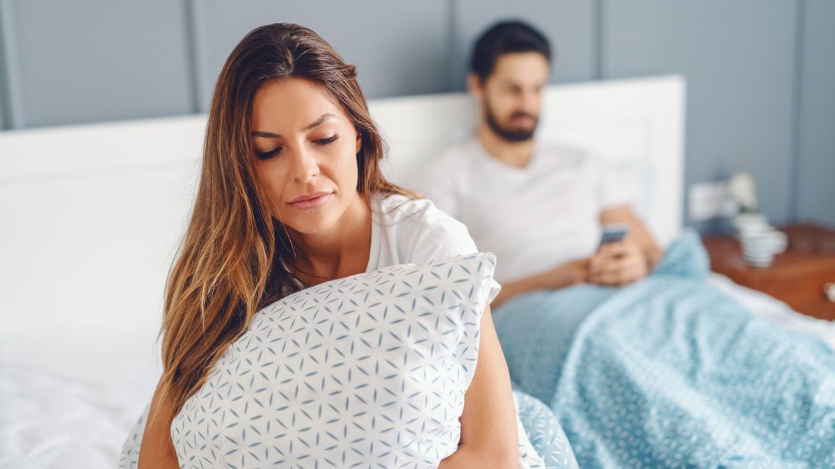 Jealous unsatisfied girlfriend holding pillow and sitting on the bed while her husband using smart phone for reading or writing message. Selective focus on woman.