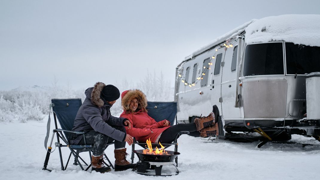 Zwei Personen in Klappstühlen sitzen vergnügt im Schnee vor einem Feuer, im Hintergrund steht ein Camper.