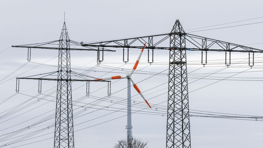 Ein Windrad steht neben dem Mast einer Hochspannungsleitung und dreht sich im Wind. Die Bundesnetzagentur will neue Netzentgelt-Rabatte für die Industrie einführen. 