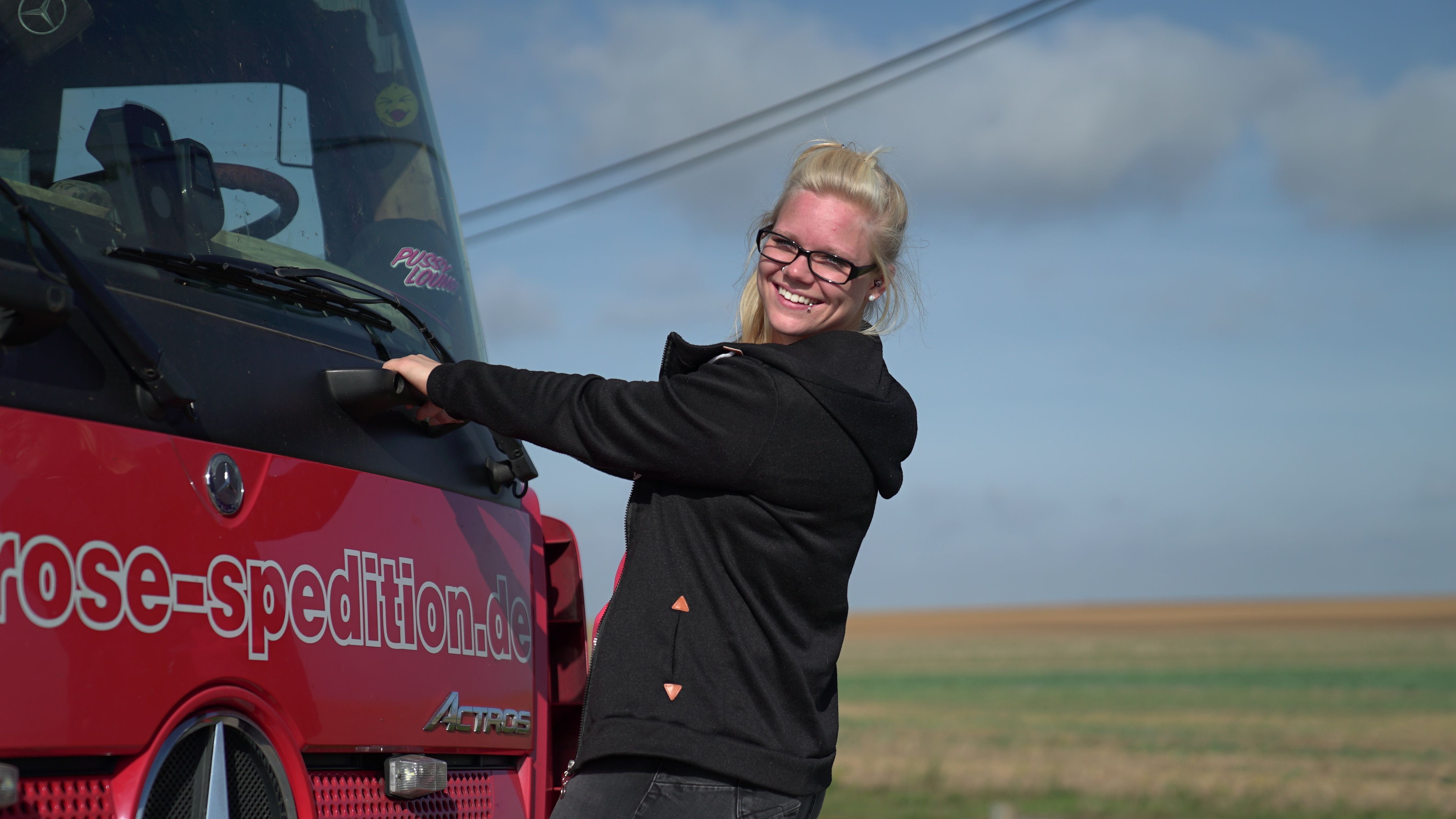 "Trucker Babe" Manu hält sich an der vorderen Fahrerkabine ihres Trucks fest und lächelt. 