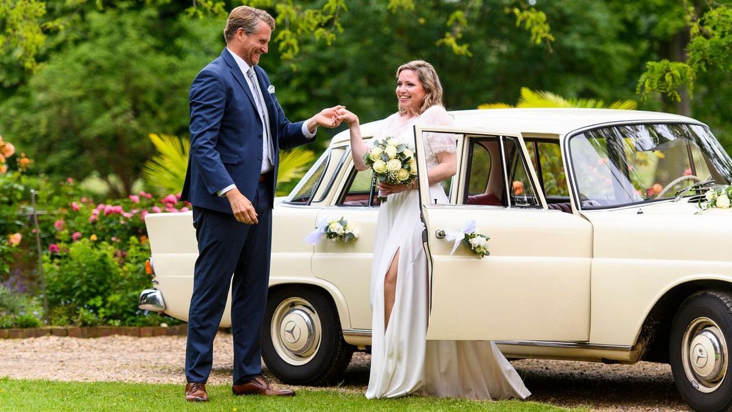 Der Trauzeuge Max reicht Sarah die Hand, als sie in ihrem traumhaften Hochzeitskleid aus dem Oldtimer steigt.