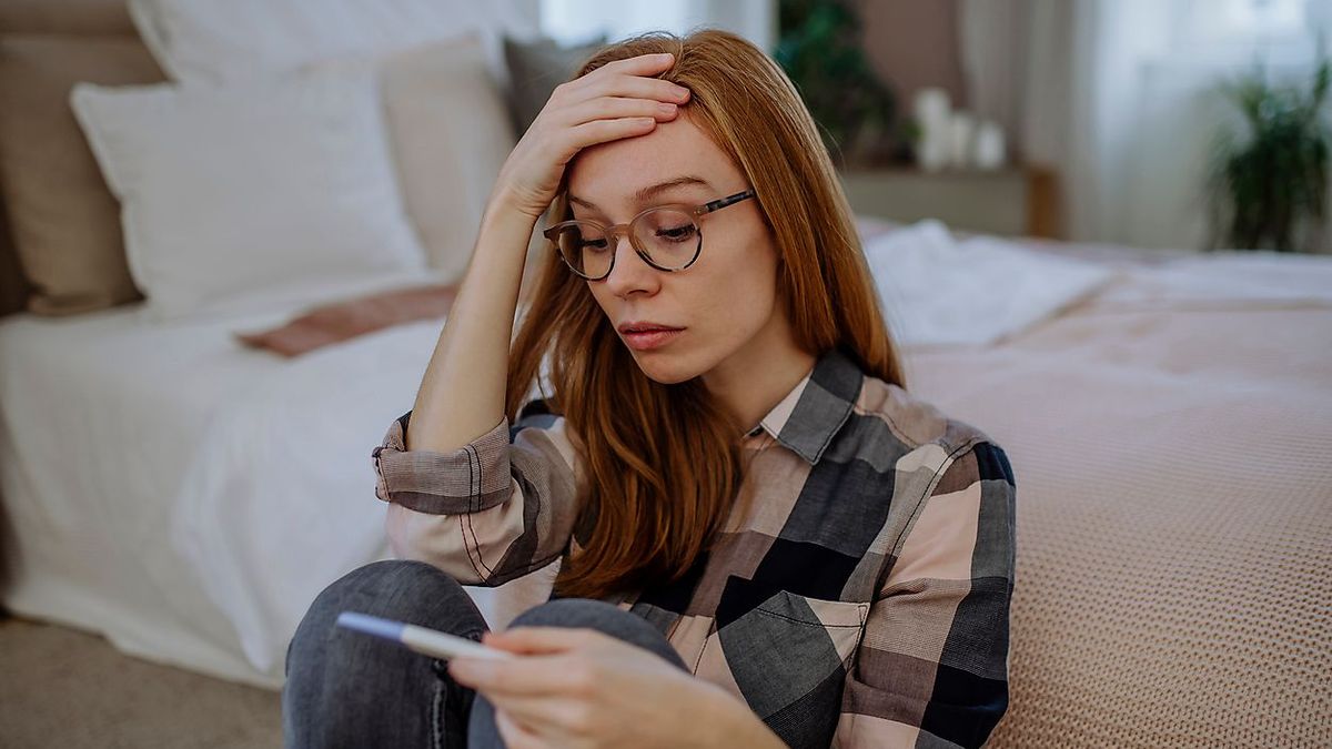 Worried woman with pregnancy testing kit sitting in front of bed at home