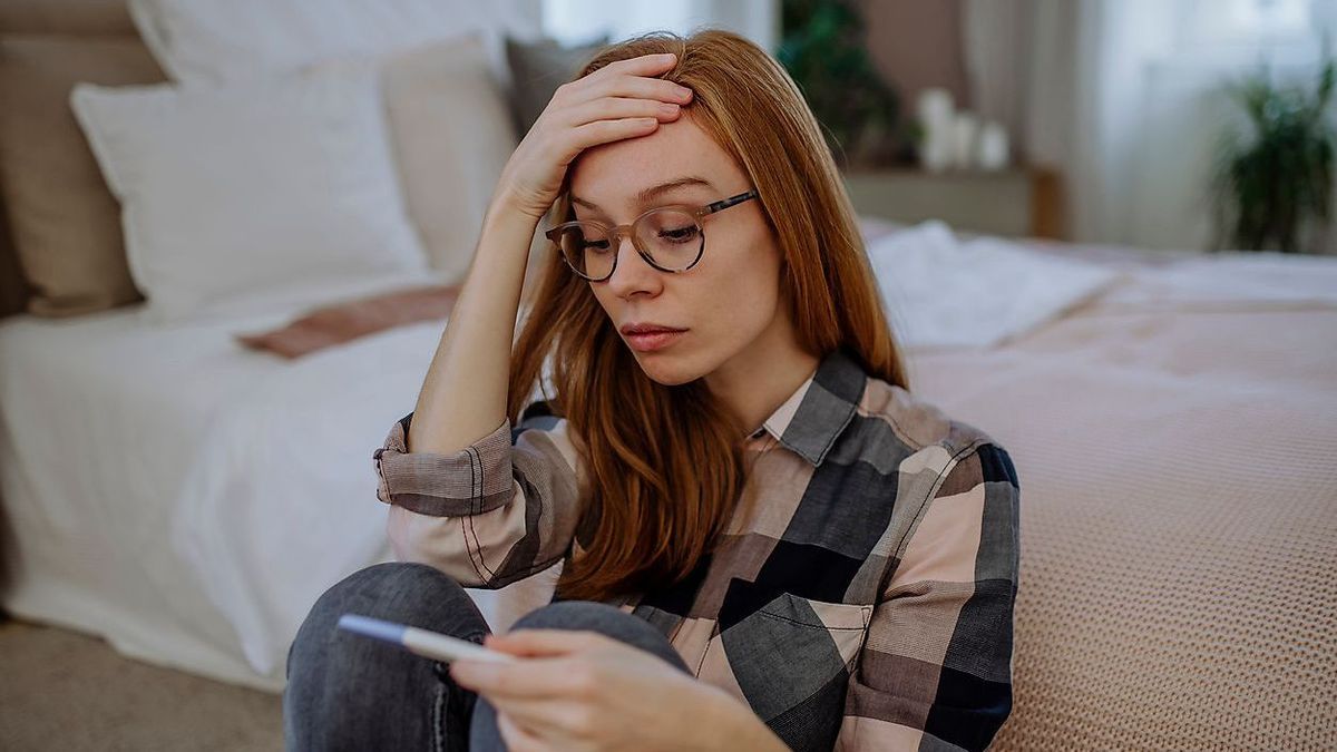 Worried woman with pregnancy testing kit sitting in front of bed at home