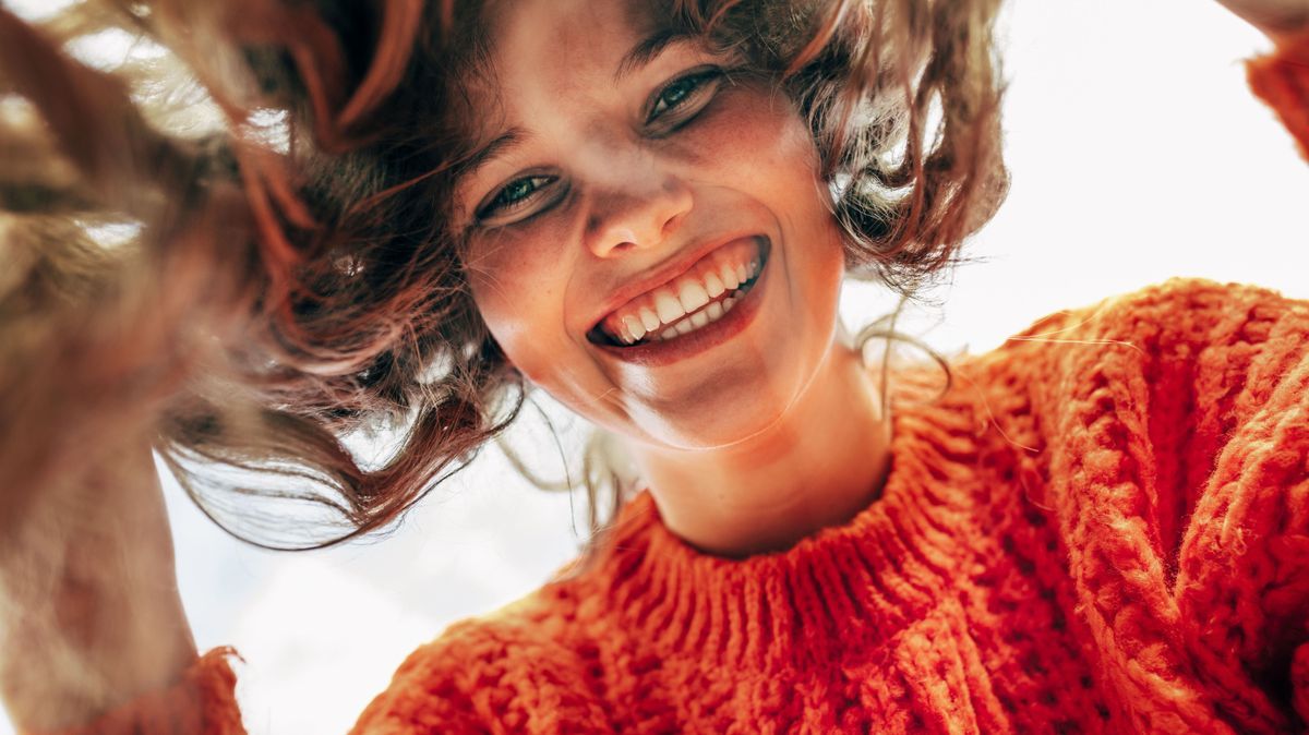 Bottom view of a candid beautiful young woman wearing a knitted orange sweater smiling broadly and looking directly to the camera outdoors. The pretty female has joyful expression, resting in the park