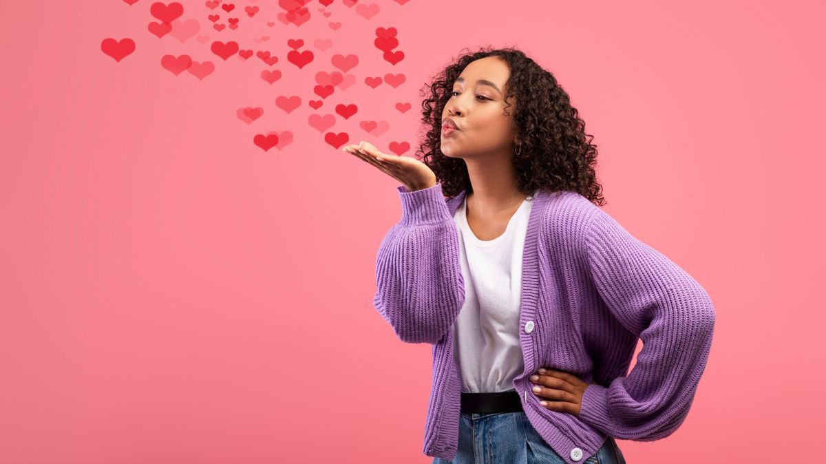 Flirty young African American woman blowing air kiss with flying red hearts over pink studio background. Valentine's Day