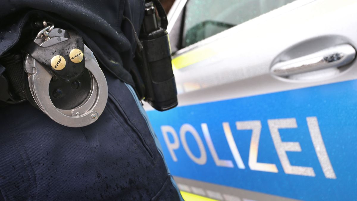 Ein Polizist mit Handschellen und einer Pistole am Gürtel steht vor einem Streifenwagen.
