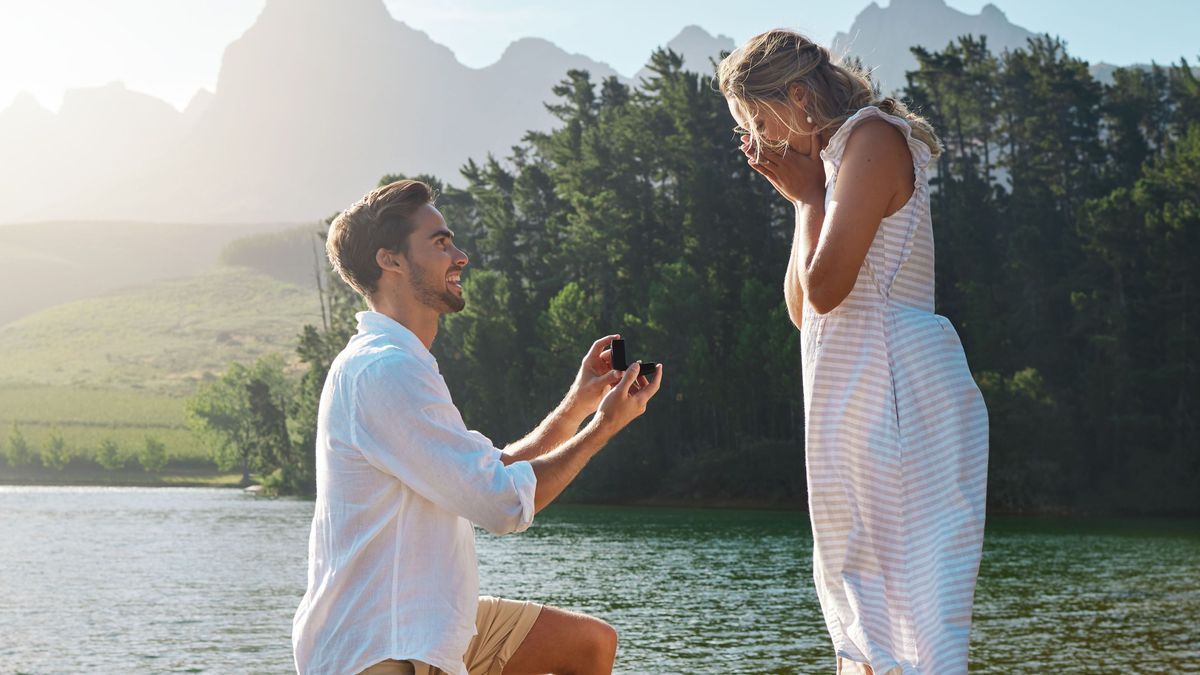 Man, woman and marriage proposal by lake on vacation with surprise, wow or happiness in sunshine. Couple, engagement and offer ring in nature for romance, love and happy on holiday in summer by water