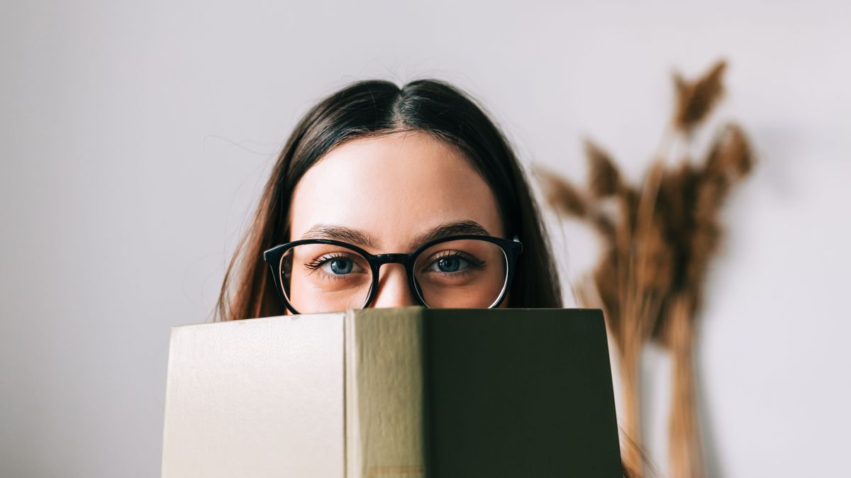 Portrait of young caucasian woman college student in eyeglasses hiding behind a book and looking at camera.