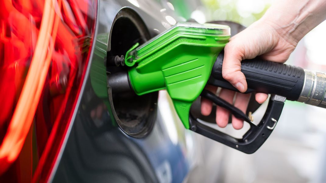Eine Frau hält an einer Tankstelle an einer Zapfsäule eine Zapfpistole in der Hand und betankt ein Auto.