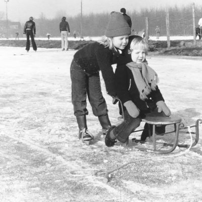 1978 vintage, seventies, retro monochrome portrait of a young girl in duffle coat on Frisian walkers pushing a boy on a retro sled on a natural ice rink.